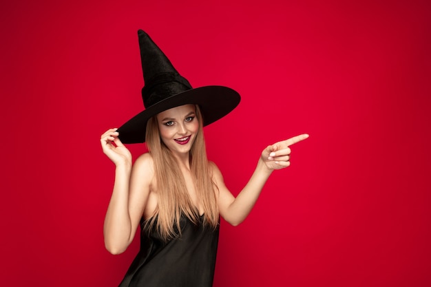 Jonge blonde vrouw in zwarte hoed en kostuum op rode achtergrond. Aantrekkelijke Kaukasische vrouwelijke model poseren. Halloween, zwarte vrijdag, cybermaandag, verkoop, herfstconcept. Kopieerruimte. Wijzen, tonen.