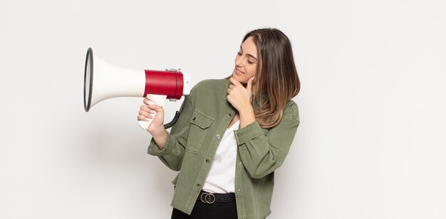 Jonge blonde vrouw lachend met een gelukkige, zelfverzekerde uitdrukking met hand op de kin, zich afvragend en opzij kijkend