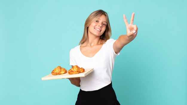 Jonge blonde vrouw lacht en ziet er gelukkig uit, gebaart overwinning of vrede en houdt een croissantblad vast