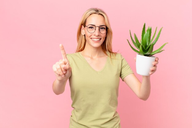 Jonge blonde vrouw lacht en ziet er vriendelijk uit, toont nummer één en houdt een cactus vast