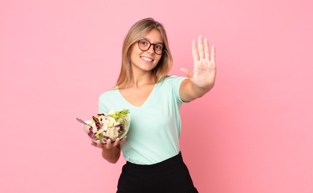 Jonge blonde vrouw lacht en ziet er vriendelijk uit, toont nummer vijf en houdt een salade vast