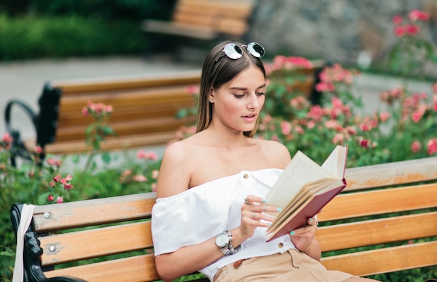 Jonge blonde vrouw lezen van een boek met enthousiasme zittend op een bankje in het park