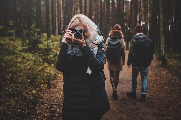 Jonge blonde vrouw maakt een foto met haar camera terwijl ze met haar vrienden in het bos wandelt.