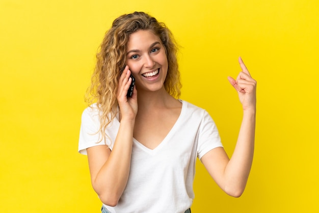 Jonge blonde vrouw met behulp van mobiele telefoon geïsoleerd op een gele achtergrond die een vinger toont en optilt in teken van het beste