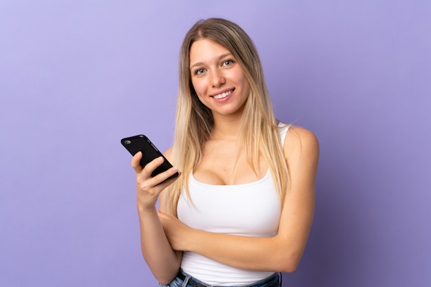 Jonge blonde vrouw met behulp van mobiele telefoon geïsoleerd op paarse muur lachen