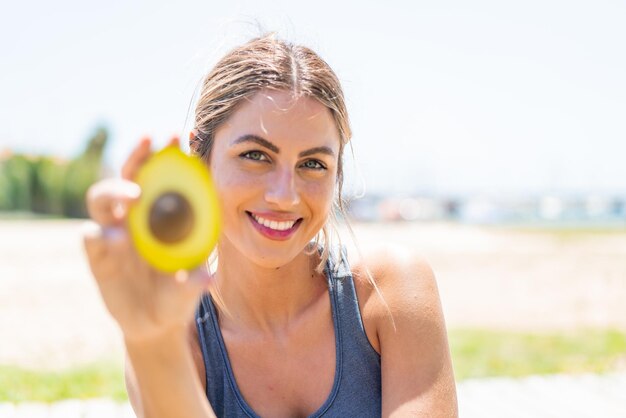 Jonge blonde vrouw met een avocado buiten met een gelukkige uitdrukking