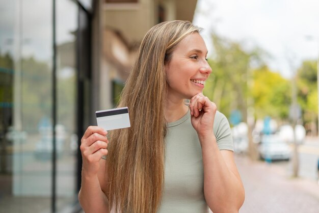 Jonge blonde vrouw met een creditcard buiten die aan een idee denkt en naar een kant kijkt