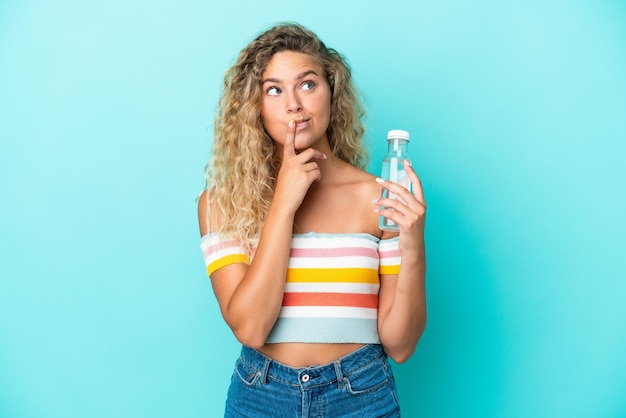 Jonge blonde vrouw met een fles water geïsoleerd op blauwe achtergrond die twijfels heeft tijdens het opzoeken