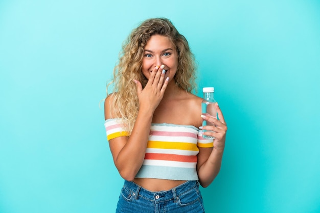 Jonge blonde vrouw met een fles water geïsoleerd op blauwe achtergrond gelukkig en glimlachend die mond bedekken met hand