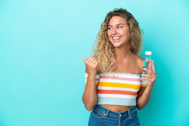 Jonge blonde vrouw met een fles water geïsoleerd op een blauwe achtergrond die naar de zijkant wijst om een product te presenteren