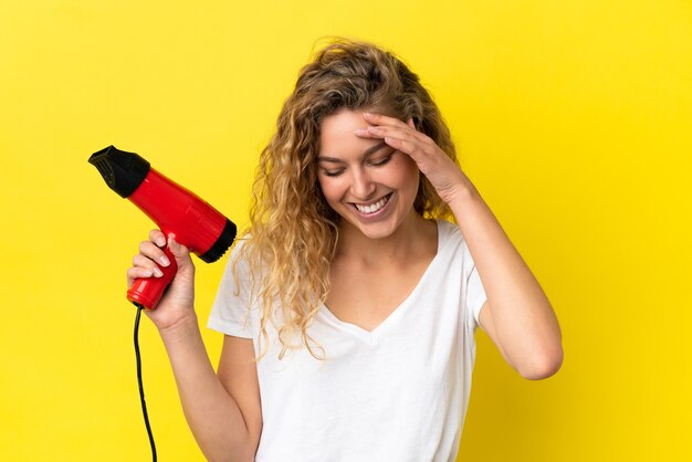 Jonge blonde vrouw met een haardroger geïsoleerd op gele achtergrond lachen