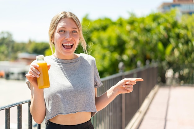 Jonge blonde vrouw met een jus d'orange in de buitenlucht verrast en wijzende vinger naar de zijkant