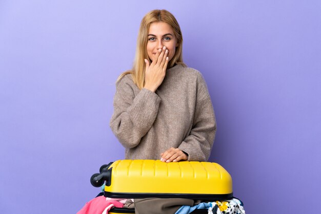 Jonge blonde vrouw met een koffer vol kleren over geïsoleerde paarse muur gelukkig en lachend die mond met de hand