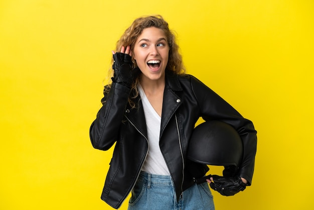 Jonge blonde vrouw met een motorhelm geïsoleerd op een gele achtergrond die naar iets luistert door de hand op het oor te leggen