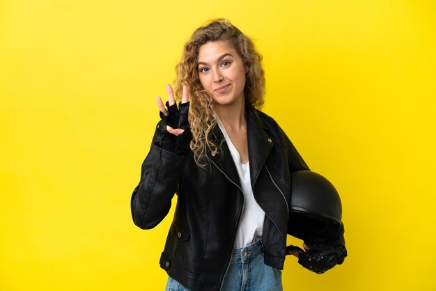Jonge blonde vrouw met een motorhelm geïsoleerd op gele achtergrond gelukkig en drie tellen met vingers