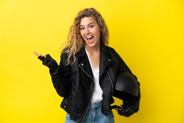 Jonge blonde vrouw met een motorhelm geïsoleerd op gele achtergrond met geschokte gezichtsuitdrukking