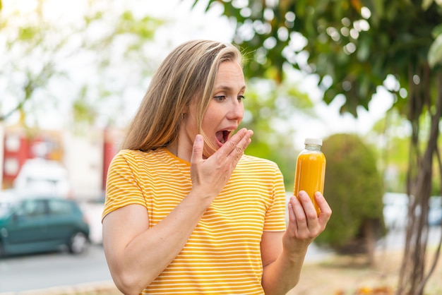 Jonge blonde vrouw met een sinaasappelsap buiten met een verbaasde en geschokte gezichtsuitdrukking