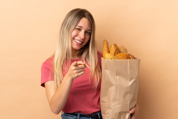 Jonge blonde vrouw met een zak vol brood geïsoleerd op beige muur wijst vinger naar je