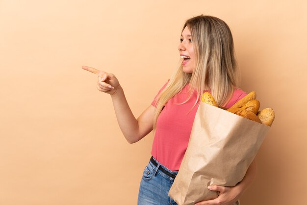 Jonge blonde vrouw met een zak vol brood geïsoleerd op beige wijzende vinger naar de zijkant