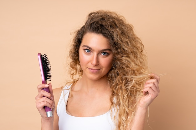 Jonge blonde vrouw met krullend haar die op beige muur met haarkam wordt geïsoleerd