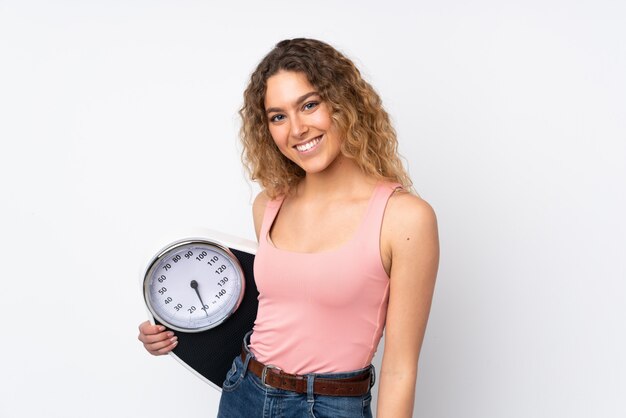 Jonge blonde vrouw met krullend haar die op witte muur met wegende machine wordt geïsoleerd