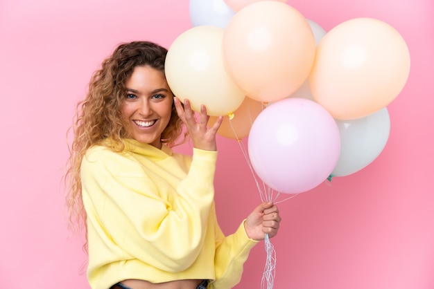 Jonge blonde vrouw met krullend haar die veel ballonnen vangen die op roze worden geïsoleerd