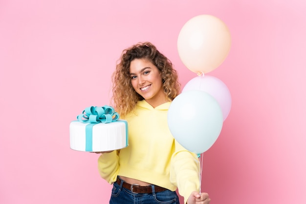 Jonge blonde vrouw met krullend haar die veel ballonnen vangt en een grote cake houdt die op roze muur wordt geïsoleerd