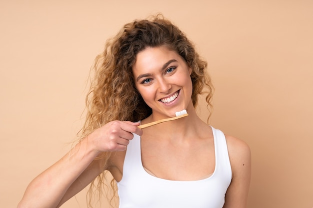 Jonge blonde vrouw met krullend haar geïsoleerd op beige met een tandenborstel en gelukkige uitdrukking