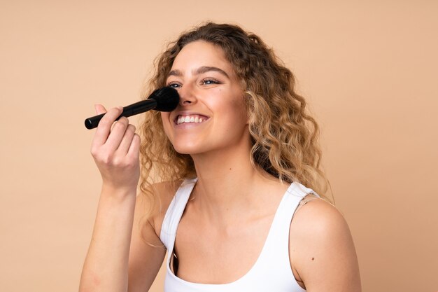 Jonge blonde vrouw met krullend haar geïsoleerd op beige muur met make-up borstel en whit gelukkige uitdrukking