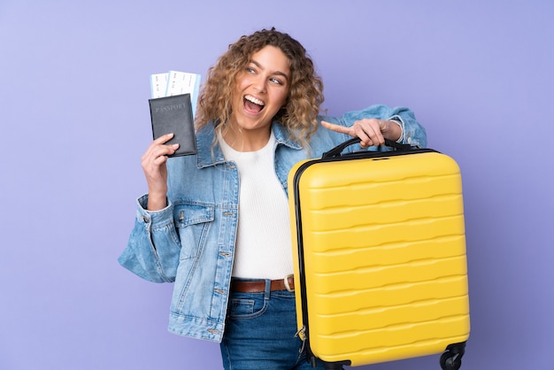 Jonge blonde vrouw met krullend haar geïsoleerd op paarse muur in vakantie met koffer en paspoort en verrast