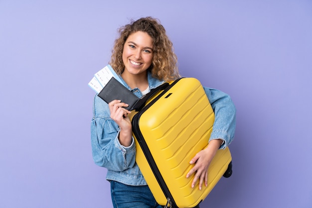 Jonge blonde vrouw met krullend haar geïsoleerd op paarse muur in vakantie met koffer en paspoort