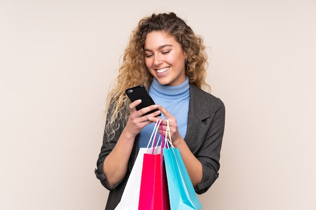 Jonge blonde vrouw met krullend haar op beige muur met boodschappentassen en het schrijven van een bericht met haar mobiele telefoon naar een vriend