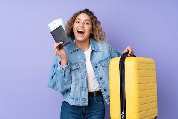 Jonge blonde vrouw met krullend haar op paarse muur in vakantie met koffer en paspoort en verrast