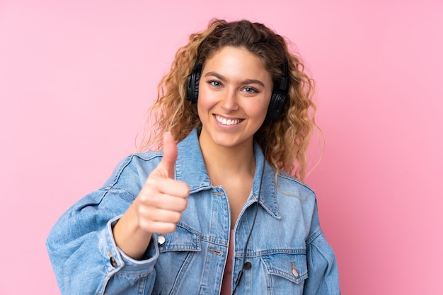 Jonge blonde vrouw met krullend haar op roze muur het luisteren muziek en met omhoog duim