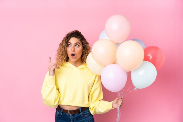 Jonge blonde vrouw met krullend haar vangen veel ballonnen geïsoleerd op roze wijzend met de wijsvinger een geweldig idee