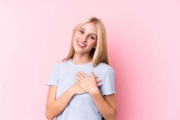 Jonge blonde vrouw op roze achtergrond heeft vriendelijke uitdrukking, palm op borst drukken. Liefde concept.