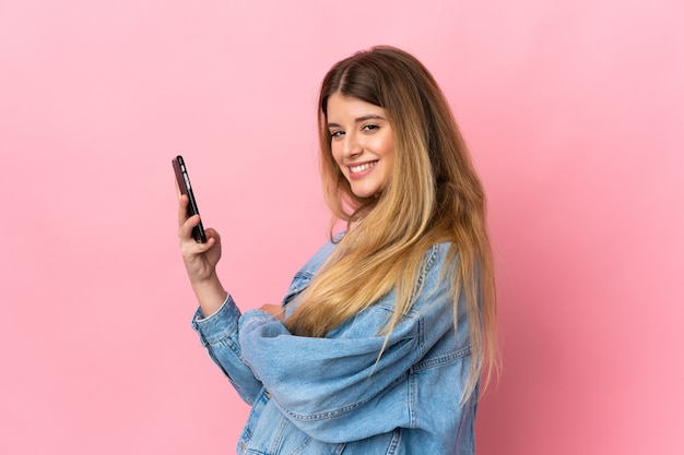 Jonge blonde vrouw over geïsoleerde muur met een mobiele telefoon en met gekruiste armen