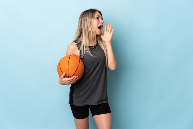 Jonge blonde vrouw speelbasketbal geïsoleerd op blauwe muur schreeuwen met wijd open mond