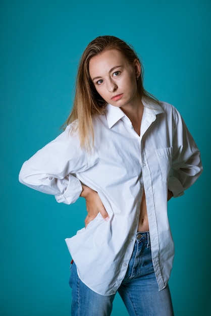 Jonge blonde vrouw van blanke etniciteit poseren in een wit overhemd en spijkerbroek op een blauwe muur