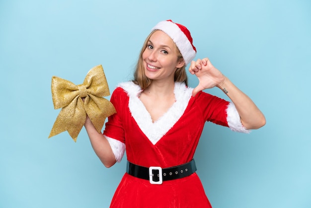 Jonge blonde vrouw verkleed als Mama Claus met een kerststrik geïsoleerd op blauwe achtergrond trots en zelfvoldaan