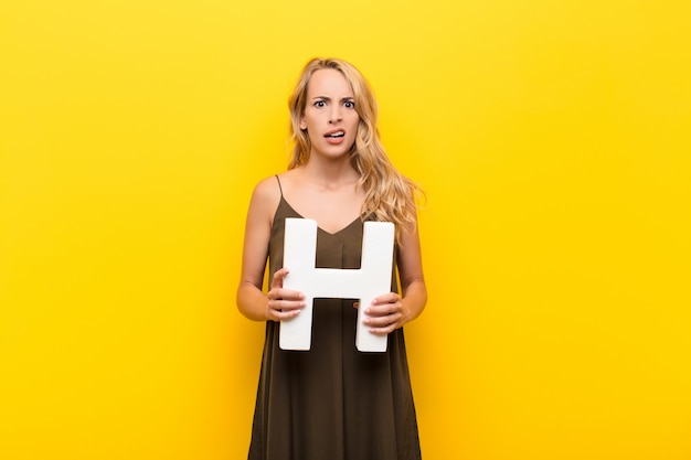 Foto jonge blonde vrouw verward, twijfelachtig, denkend, met de letter h van het alfabet om een woord of een zin te vormen.