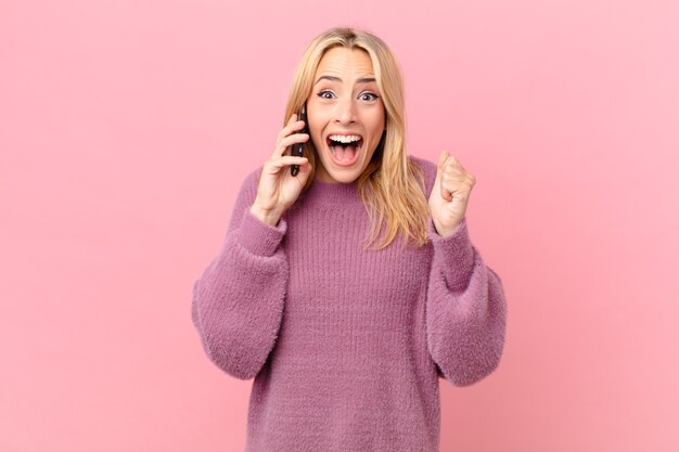 Jonge blonde vrouw voelt zich geschokt, lacht en viert succes en praat met een smartphone