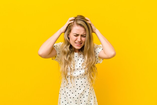 Jonge blonde vrouw voelt zich gestrest en gefrustreerd, steekt haar handen op, voelt zich moe, ongelukkig en met migraine over de gele muur