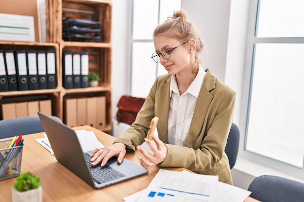 Jonge blonde vrouw zakelijke werknemer met behulp van laptop eten granen bar op kantoor