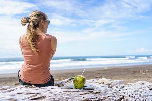 Jonge blonde vrouw zittend op het strand met een open kokosnoot met een rietje. Jaco strand in Costa Rica