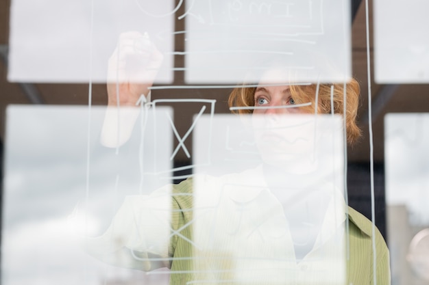 Jonge blonde vrouwelijke softwareontwikkelaar die zich concentreert op het werk over een nieuw project terwijl ze voor een transparant bord met schetsen staat