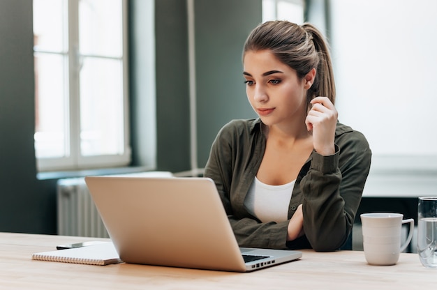 Jonge blonde vrouwelijke student die aan laptop werkt die bij het lezen van het scherm concentreert zich.