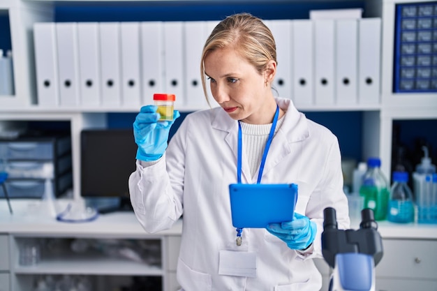 Jonge blonde vrouwelijke wetenschapper die touchpad gebruikt en urinereageerbuis vasthoudt in het laboratorium