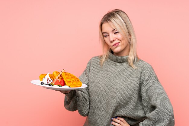 Jonge blonde vrouwenholdingswafels over geïsoleerde roze muur met droevige uitdrukking