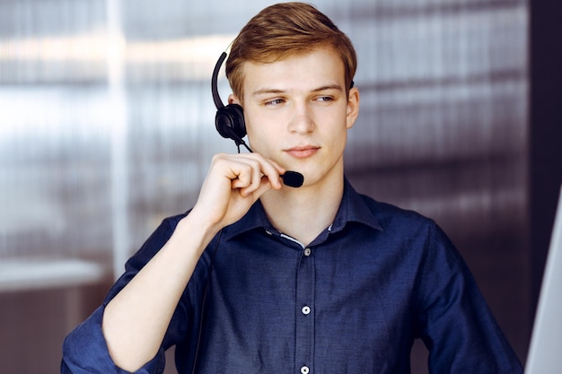 Jonge blonde zakenman die hoofdtelefoon en computer met behulp van op het werk. Opstarten betekent hard werken en geen tijd hebben om succes te behalen